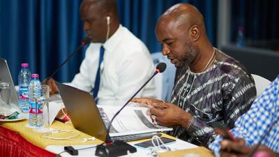 Man in West African print shirt sits behind microphone at conference table