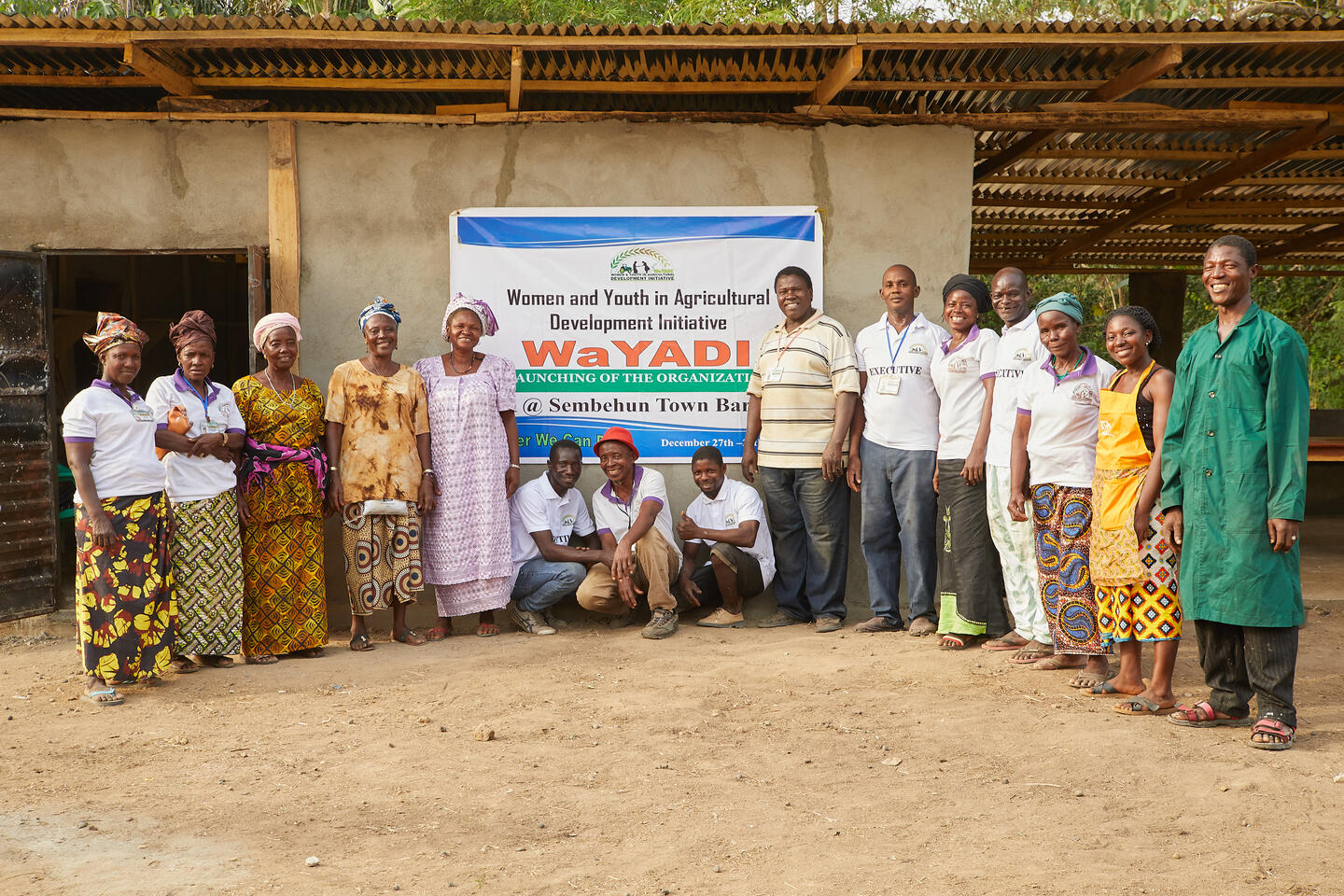 Group of cassava farmers stands next to WaYADI sign on dusty field outside building