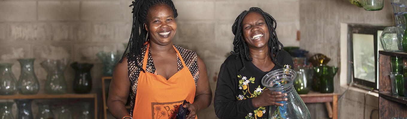 Two women of African descent with big smiles, one of them wearing an orange apron. There are low shelves of glass vases behind them along a brick wall, and one woman is holding a large glass vase