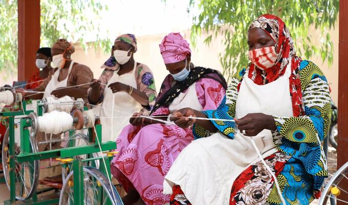 Five Malian women, seated outside under a covered porch, wearing fabric facemasks, and weaving white thread using simple machines.