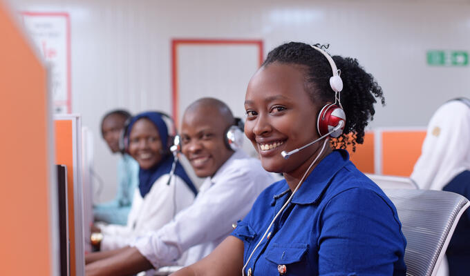 4 call centre people wearing headsets sitting in a row in West Africa, woman in front wearing blue and a big smile