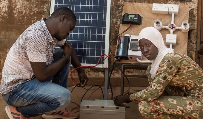 Man and woman crouching with a small solar panel and its equipment in center of picture.
