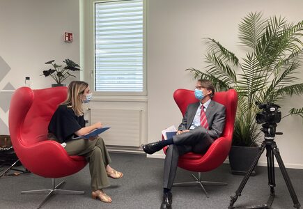 Woman with clipboard interviewing man, both seated in red chairs, camera to right side of image.