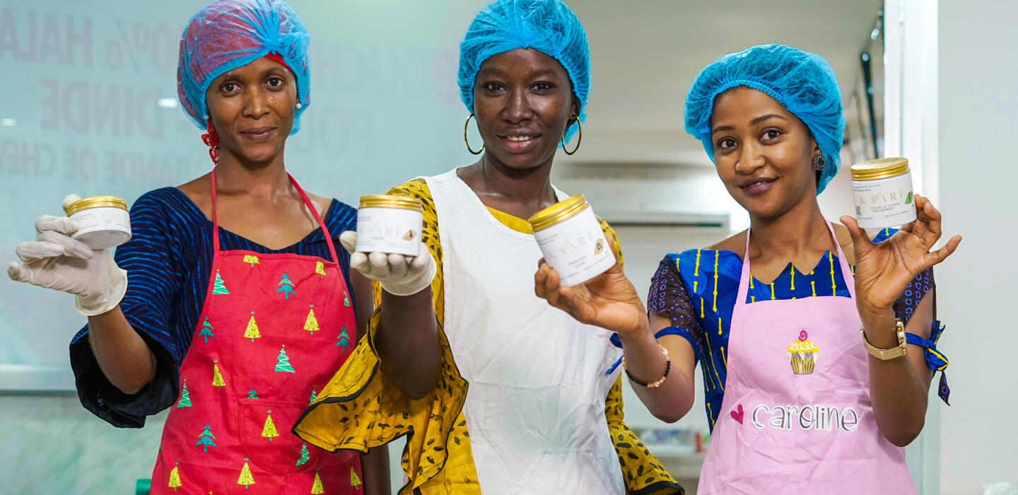 Three women wearing blue hairnets and colourful aprons holding shea products.