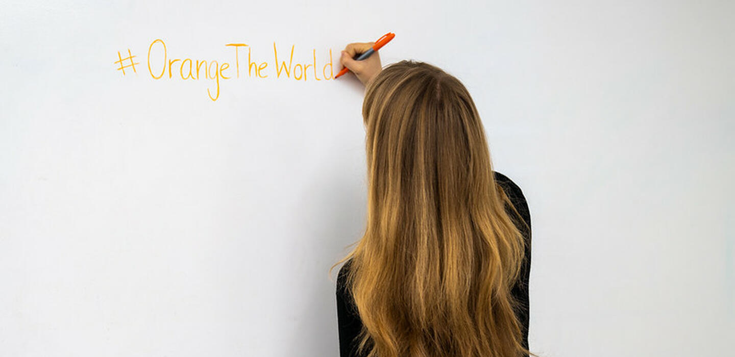 Woman with long hair with back to camera writing "#Orange The World" on a white wall in orange