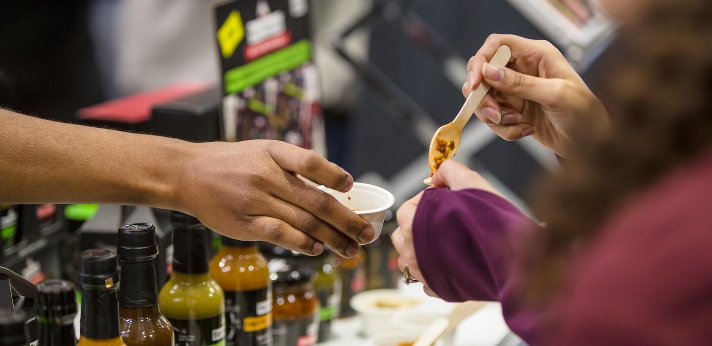 Woman holds out small bowl with samples of chilli sauce for a customer holding a spoon.