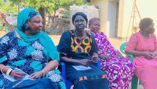 Three South Sudanese women in brightly coloured clothing sit outside