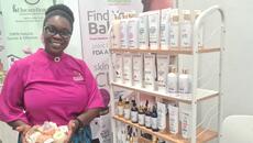 Ghanian woman in pink t-shirt holds a tray of soaps next to a stand of beauty products.  